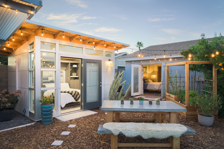 A small ADU house color white with exterior lights color yellow, a white table and small garden